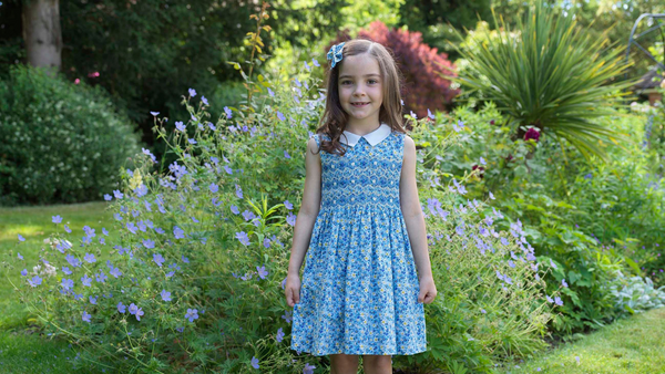 Young girl in Rachel Riley Spring Floral Smocked Dress. Blue floral print with contrasting ivory white collar, hand embroidered chest.