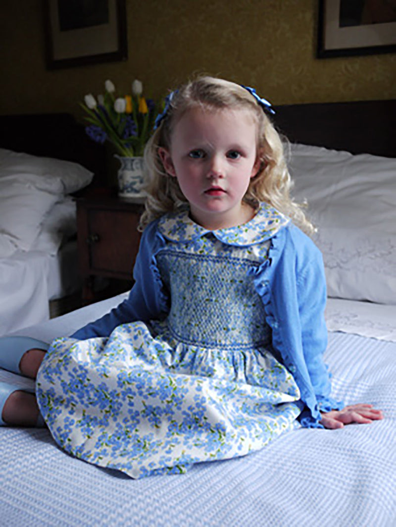 Girl in blue floral dress with smocked bodice, peter pan collar and gathered skirt, styled with blue frill cardigan.
