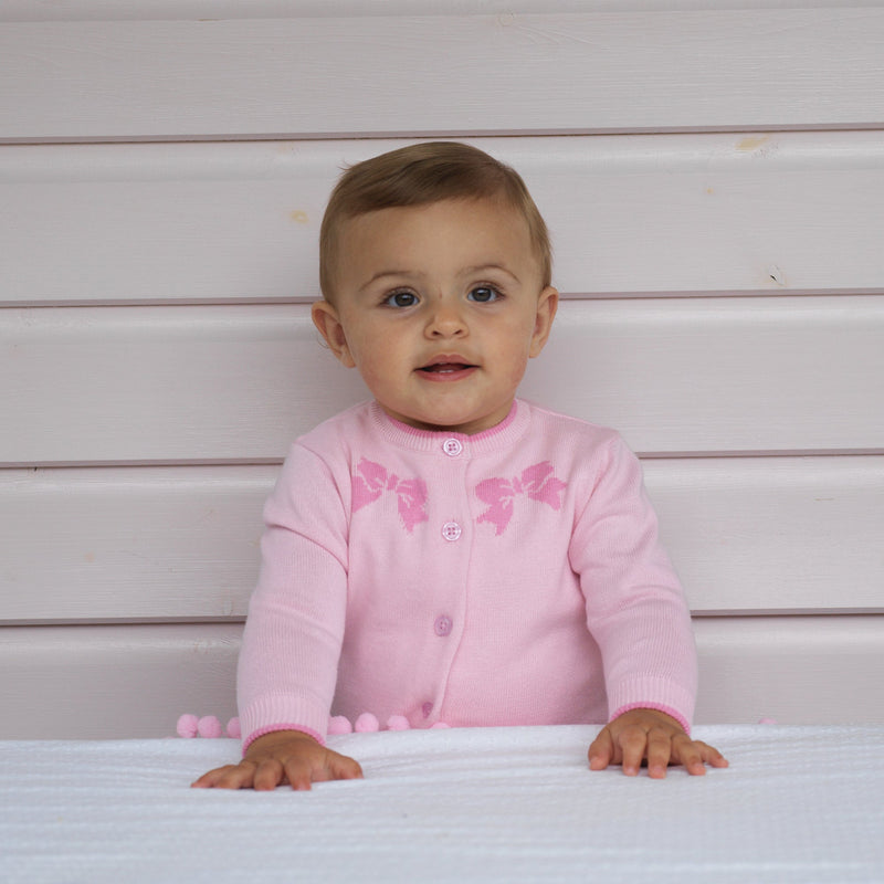 Baby girl wearing knitted pink cardigan with intarsia bow design on the chest area and trousers set.