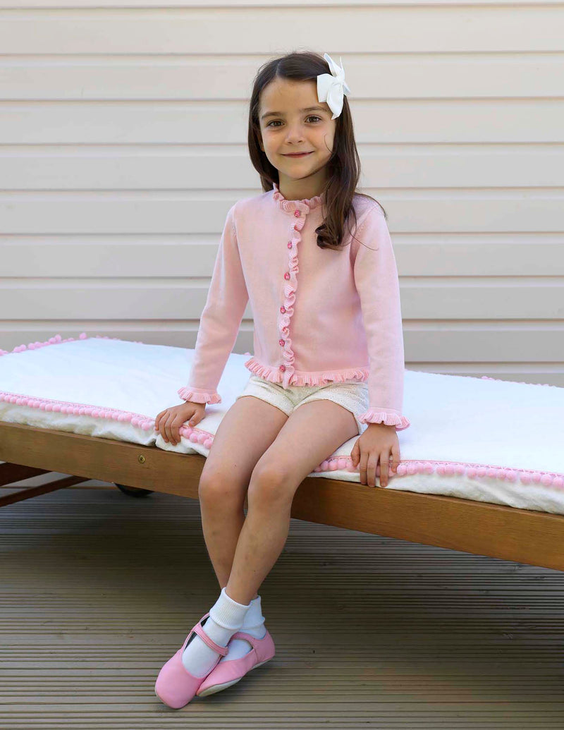 Girl in pink cotton cardigan trimmed with frills at front, neck and sleeves and jewel buttons.