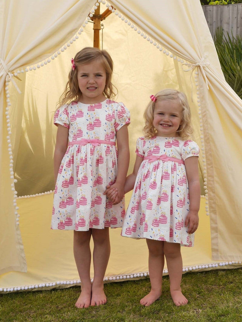 Girls in matching party dress with a pink birthday cake print and pink ribbon and bow at waist.