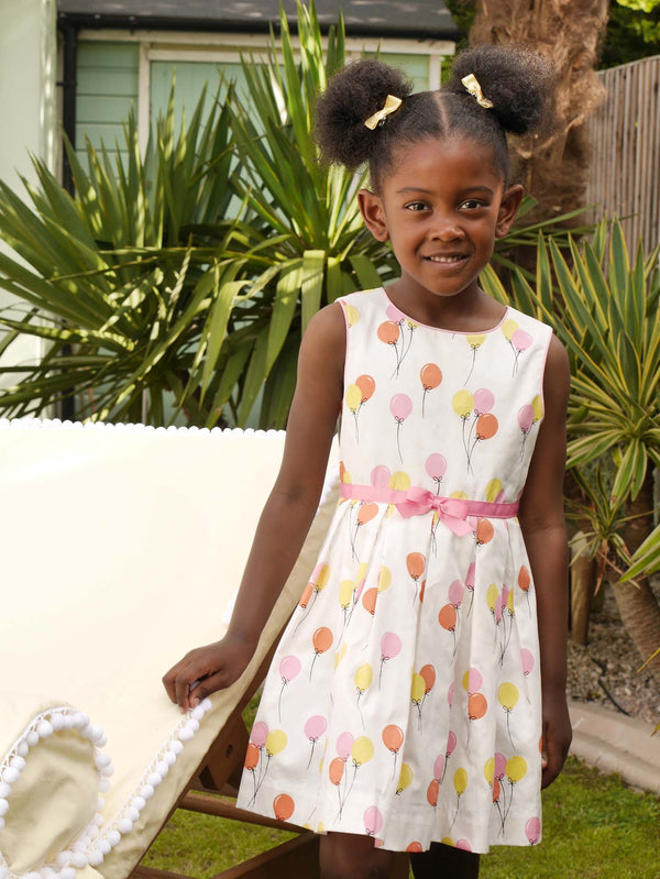 Girl in sleeveless pleated dress with pink and yellow balloon print and pink ribbon and bow at the waist.