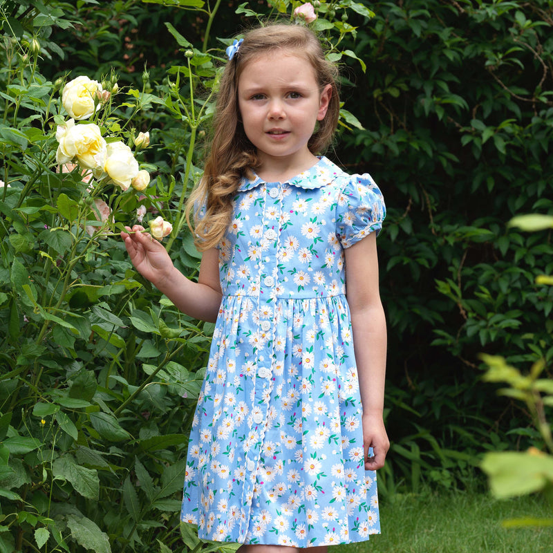 Girl in button-front dress with ivory blossom daisy print on soft blue base, contrasting ricrac trim and buttons.