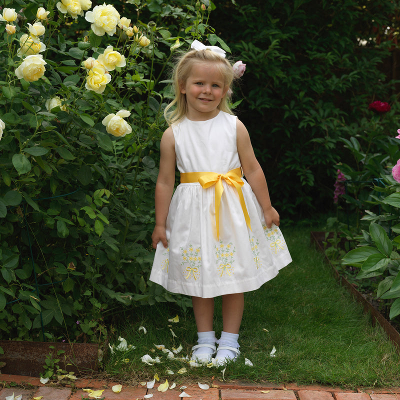 Girl in sleeveless ivory dress with yellow and green mimosa embroidery at skirt and ribbon to tie