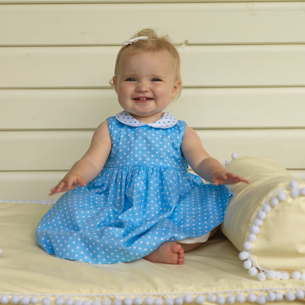 Baby girl in blue polka dot print dress, sleeveless with contrasting peter pan collar and sash tie.