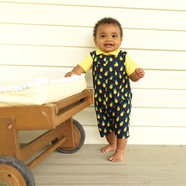 Baby boy wearing navy dungarees with pineapple print and yellow polo shirt.