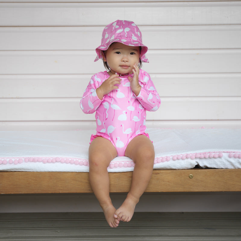 Baby wearing one piece rash guard with pink flamingo print and matching flamingo print sunhat.