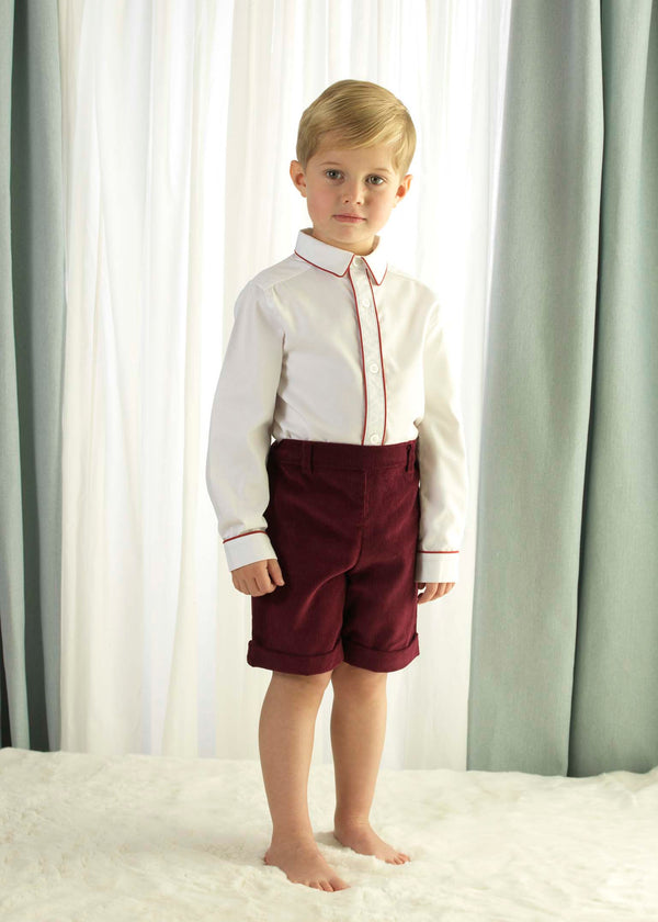 Boy wearing white pique shirt, trimmed with burgundy pipping and matching red corduroy shorts