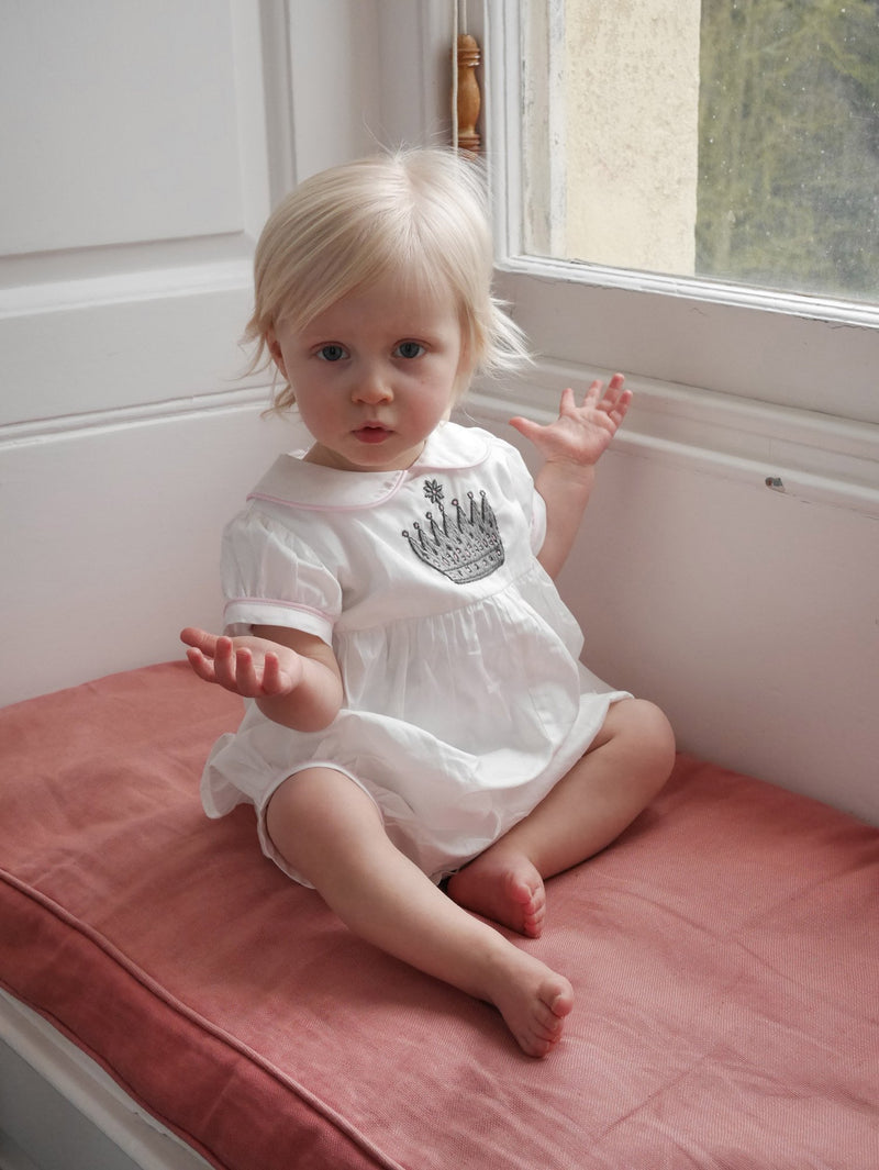 Baby girl in ivory babysuit with crown embroidered motif on chest and pink piping at collar and cuffs