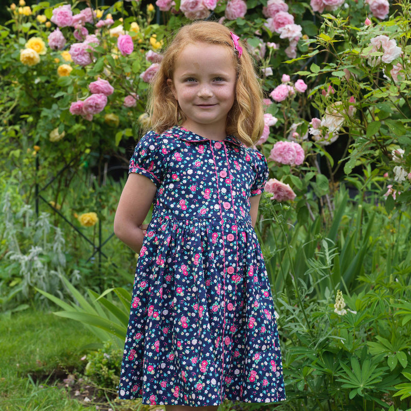 Girl in navy button-front dress with floral print, puff sleeves and pink ricrac trims and buttons.