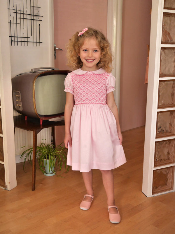 Girl in pink dress with smocked bodice, embroidered with bows, peter pan collar and gathered skirt.