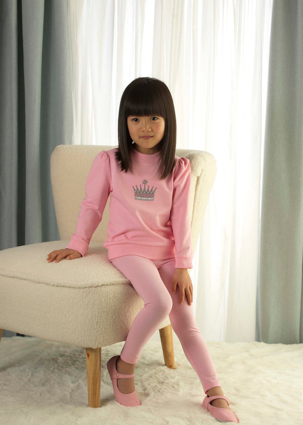 Girl in pink jersey sweatshirt with embroidered crown motif at the chest and matching leggings.