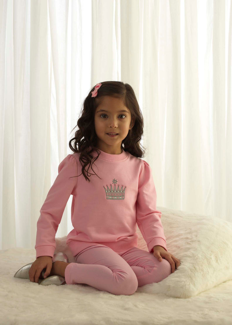 Girl in pink jersey sweatshirt with embroidered crown motif at the chest and matching leggings.