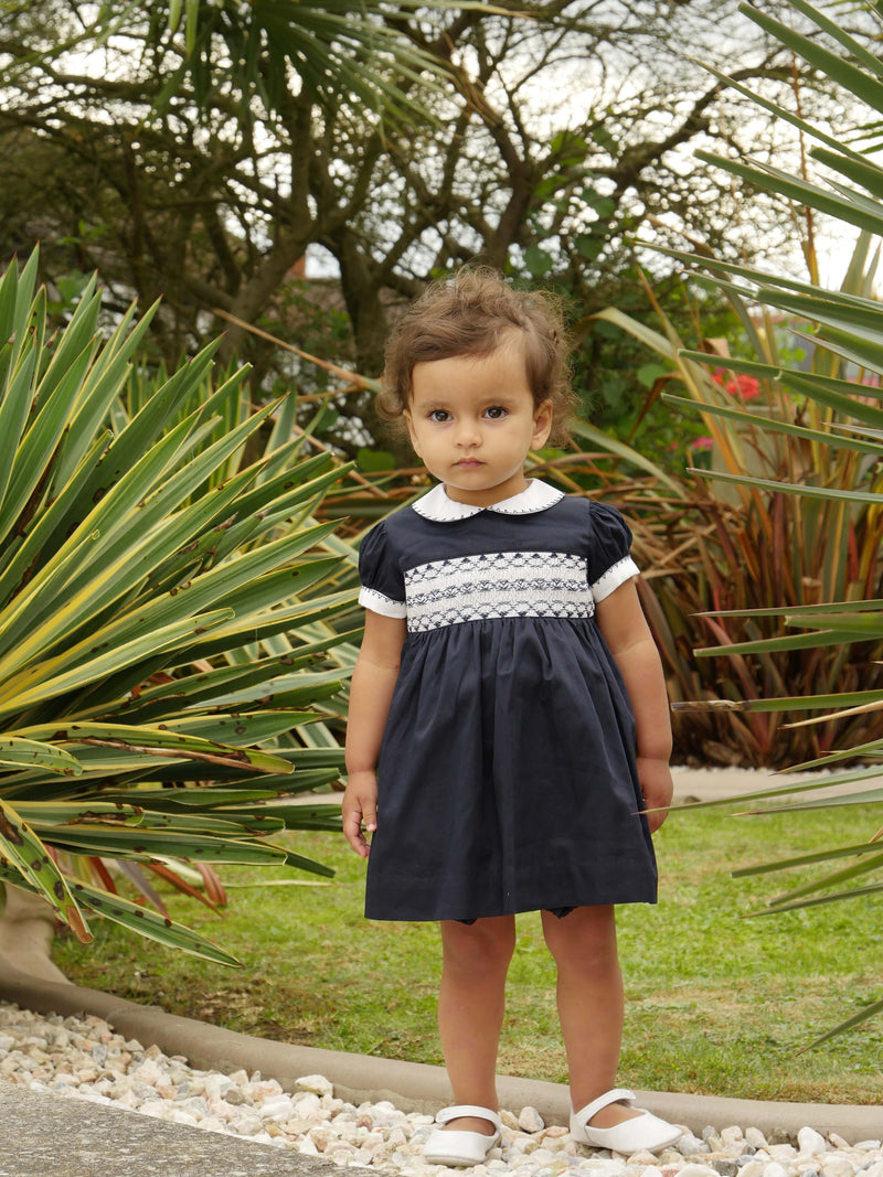 Baby wearing navy classic smocked dress, styles with matching hairbow and slippers.