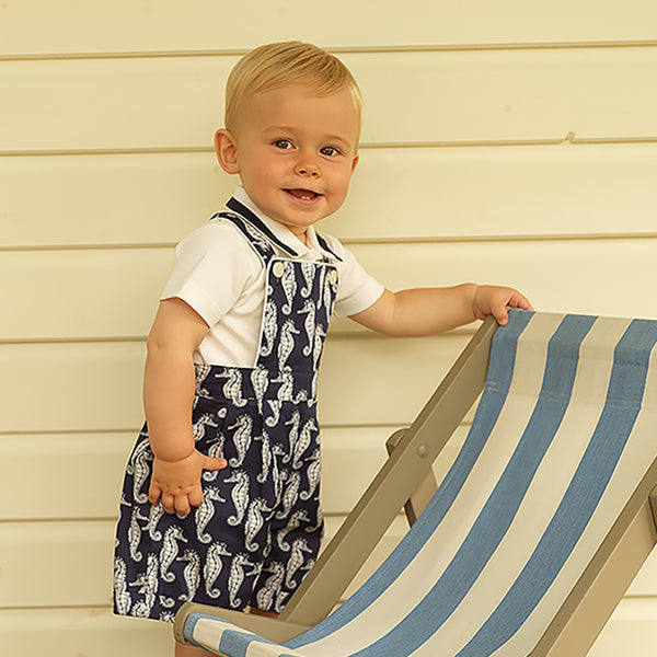 Baby boy wearing seahorse print navy dungarees styled with ivory jersey short sleeved polo shirt